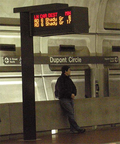 This photo shows a man waiting at the Metro Station in Dupont Circle. Mounted on a post to the left of the man is an electronic sign that provides the estimated times of arrival for approaching trains.