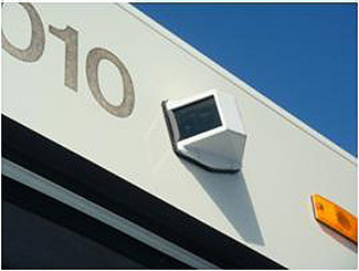 This photo is an example of an exterior video surveillance camera. This square camera is in the center of the photograph and mounted to the side of a white metal structure.
