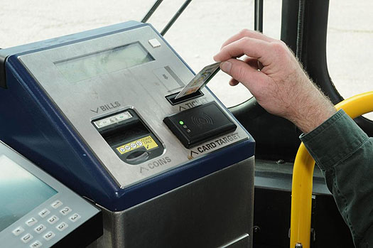 This is a photograph of an automated farebox. In this photo there is an electronic box with a slanted top facing the passenger. At the top of the slanted face is a digital display. On the left is an area to make payment and deposit cash. On the left is a person sliding a card into a card reader. Below the card reader is a rectangular card target sensor.