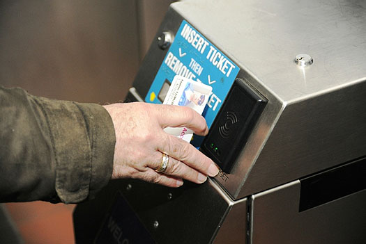 This is a photograph of an automated faregate. In the photo is a metal box. On the top portion of the box, there is a card reader on the right side of the passenger facing surface. On the left is a blue sign that says: Insert Ticket then Remove Ticket. A person wearing a brown jacket is holding up their smartcard to the card reader.