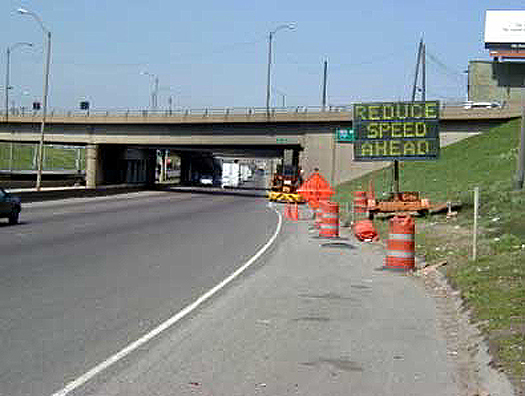 This is a photograph of a Portable Dynamic Message sign. This sign is placed on the side of the road in front of a road construction site. The message on the board reads: Reduce Speed Ahead.