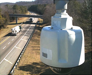 This is a photograph of trucks passing underneath a dome camera mounted high above a two-lane road. This camera is in a standard enclosure, and the camera dome is located at the bottom.