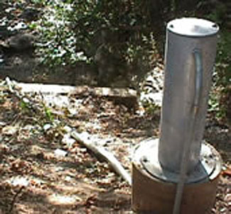 This is a photograph of a stilling well. It is a tall metal cylinder affixed to a concrete cylinder in a wooded area with dirt ground.
