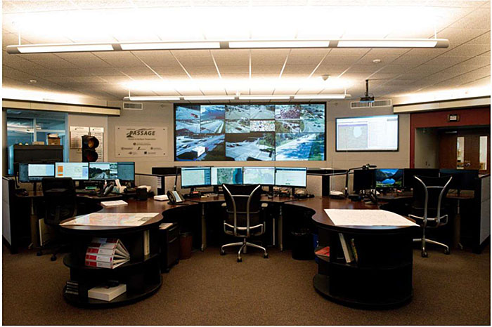 Photo showing an example county transportation management center in Lake County Illinois with a large desk and numerous computer screens and display feeds from cameras.