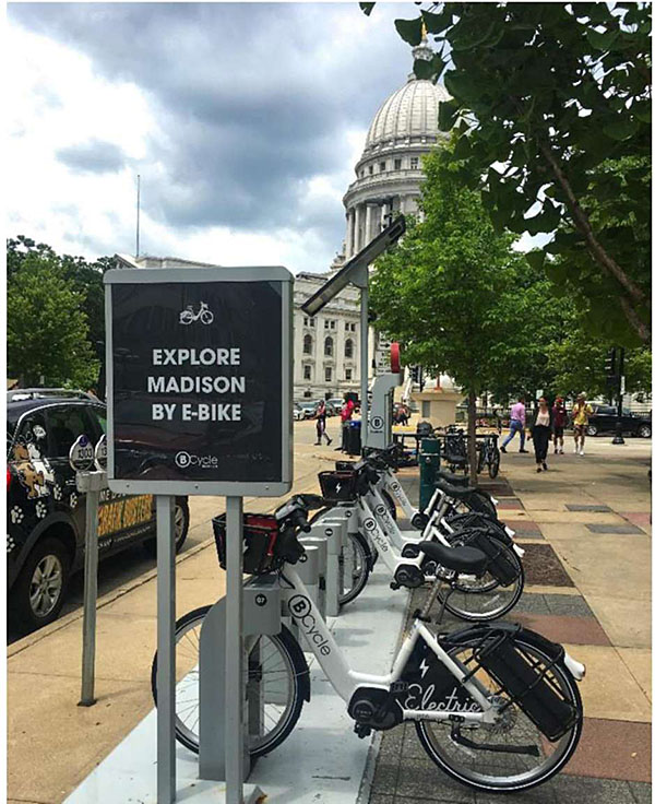 Photo of E-Bike sharing program in Madison Wisconsin, with a row of E-bikes in a rack along the street with a sign next to them that says Explore Madison by E-bike.
