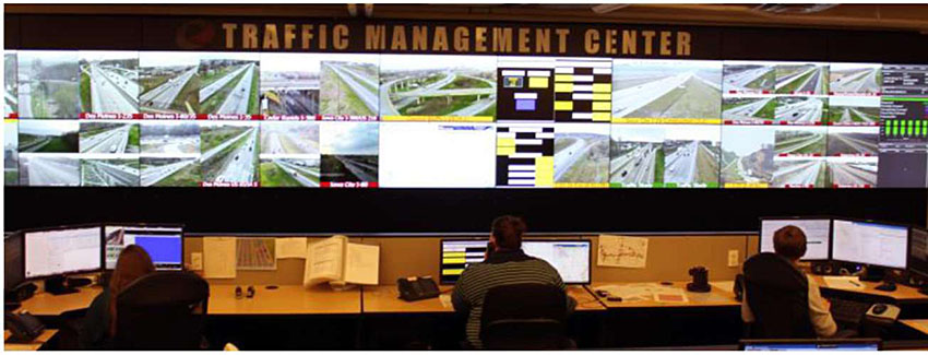 Photo showing an example state DOT traffic management center in Iowa with three operators at a series of desks with several computer screens and a large panel of computer screens and camera displays on the wall in the background.