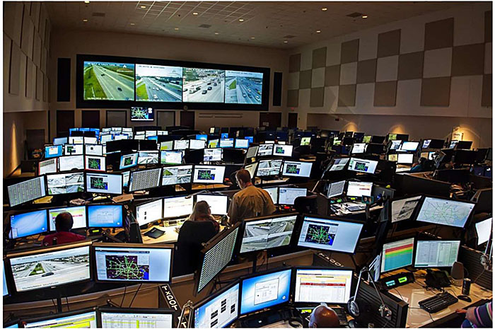 Photo of an example multi-agency transportation management center in Houston showing a large room with several rows of desks, all with numerous computer screens for a larger number of operators, with a large panel of displays on the wall in the background.