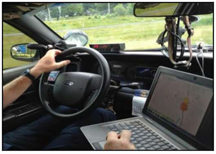 Photo of interior of law enforcement vehicle showing a laptop computer in the vehicle as the officer is parked along the roadside. The laptop shows a map.