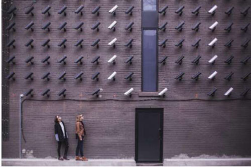 The slide shows the wall of a building with a 6 by 14 array of CCTV cameras pointing down at two women who are on the sidewalk looking back at the cameras.