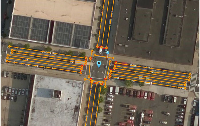 The slide entitled "What are the mandatory elements of a MAP message" has a graphic that shows an example aerial view of an intersection in New York City. There is a one-way main street from right to left with a total of six lanes. There is a one-way side street going down with a total of three lanes. Each lane is indicated with an orange line and a number that represents the lane identifier. There are nodes visible at the start of each lane at the stopbar of the adjacent intersection to the stopbar of that lane at the intersection. In the center of the intersection is a blue bubble that represents the reference point. There is also a crosswalk at each side of the intersection. Each crosswalk is indicated with an orange line and a node. There also red squares on each side of the intersection, indicating the locations of stop bars. There is a blue pin is at the traffic pole at the upper right corner of the intersection that is used as a survey point used by a tool that can be used to check for positional accuracy.