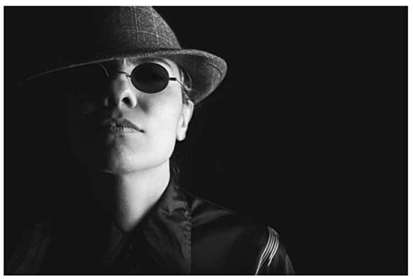 This slide has a rectangular photograph of a woman from the shoulders up. The picture is mostly dark with the light illuminating the left side of her face. She is wearing a fedora hat, dark glasses, and what appears to be a buttoned up jacket. The picture is to the lower right of the slide text.