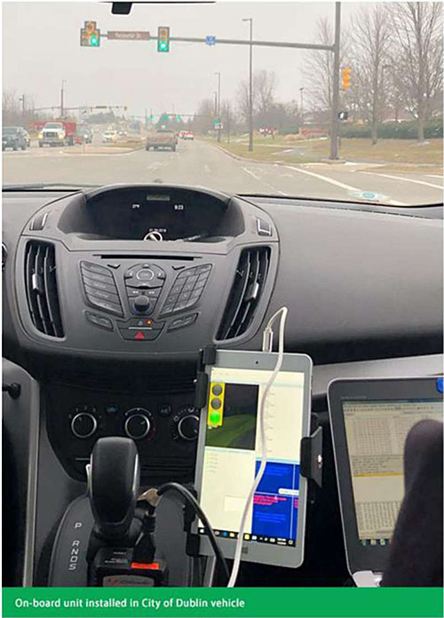 Photo shows a moving vehicle equipped with OBU near the center console of the car at an iintersection with a green light, showing the green light on the OBU screen. Label says On-board unit installed in City of Dublin vehicle.
