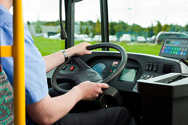 A photo of a seated bus driver holding the steering wheel and a mobile data terminal on top of the dashboard.