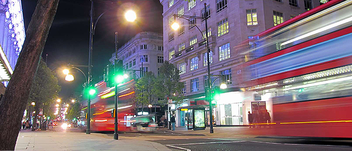 A photograph of an urban city street with 3 traffic signal heads and a pedestrian crossing. There are two red transit vehicles, both buses were captured while moving and shows a motion blur following it, which makes the buses look like they were traveling at a high velocity. Image Source: thinkstock.com 