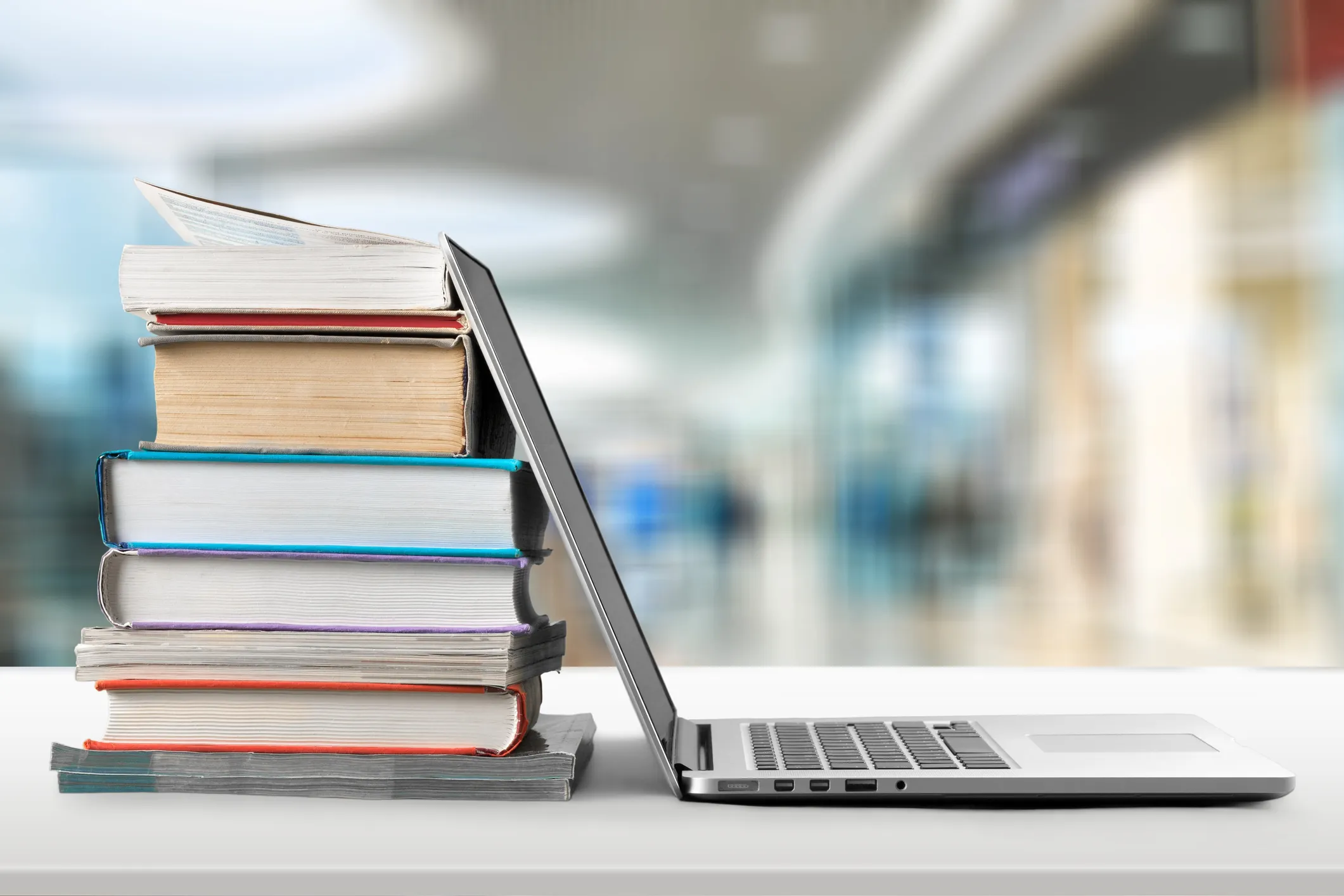 A stack of colorful books leaning against a laptop on a desk with a blurred library background.