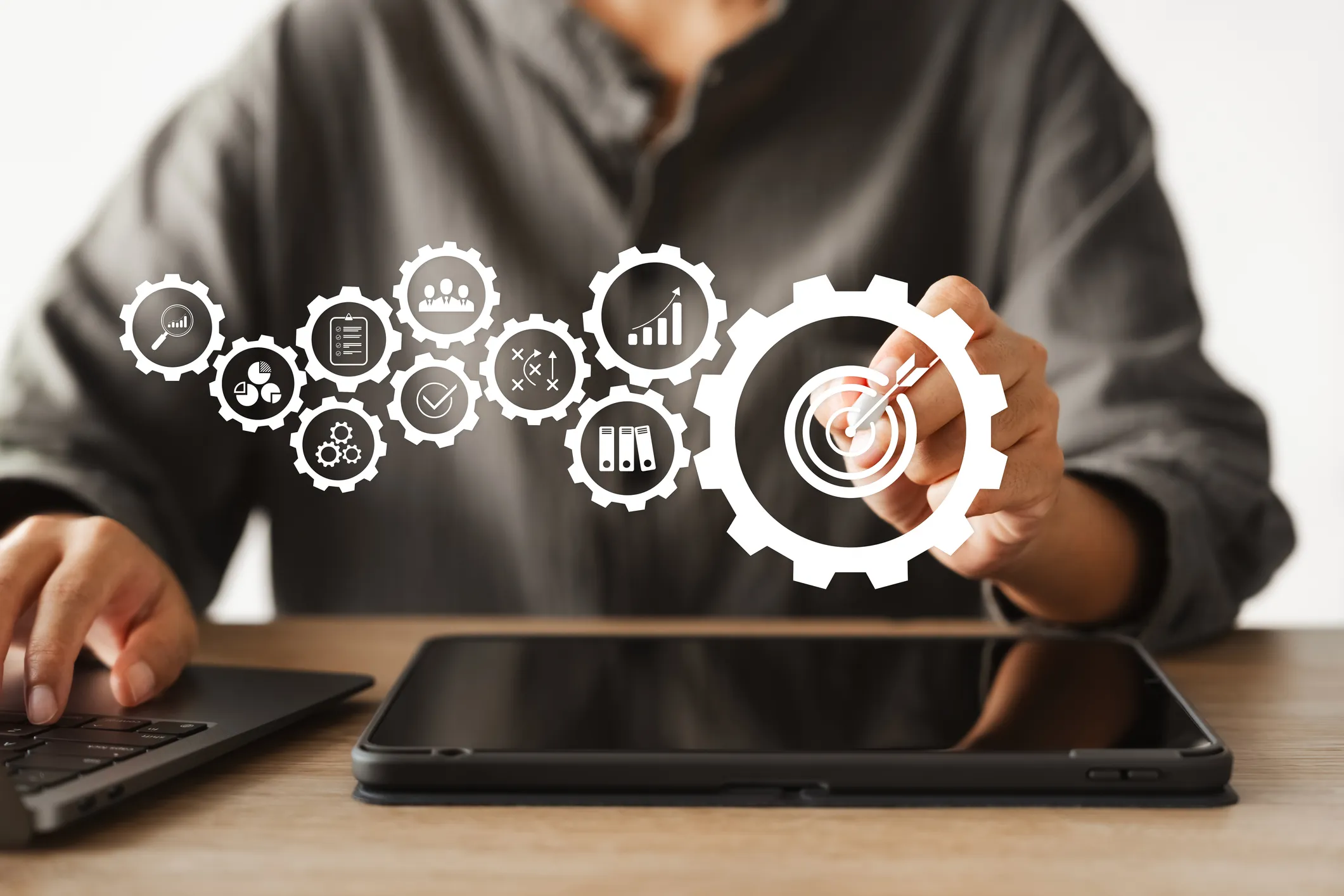 A professional in business attire interacts with a tablet, manipulating floating gears with various business icons (analytics, charts, team management, checkmarks, strategy).
