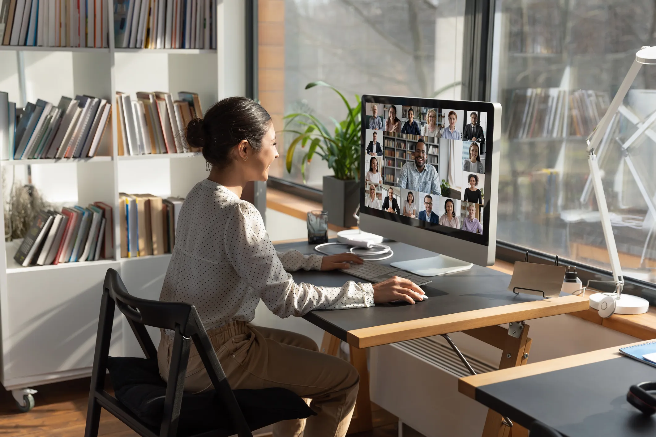 A woman sits at a desk with bookshelves behind her, viewing a video conference call with multiple participants displayed on a monitor in a bright office setting.