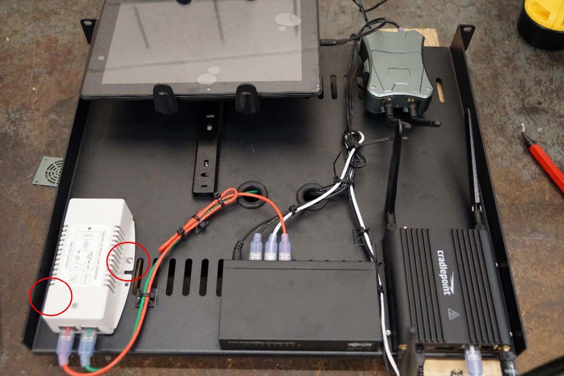 This photo demonstrates the proper placement of a Power over Ethernet (PoE) injector on a component shelf. The placement of the 6-32 screws is circled in the image. The PoE injector is placed below the tablet and to the left of the network switch.