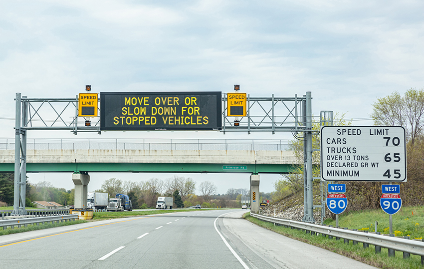 a photo of an overhead dynamic messaging sign (Move Over or Slow Down for Stopped Vehicles) which is flanked by two dynamic speed limit signs. On the side of the highway are stationary speed limit signs.